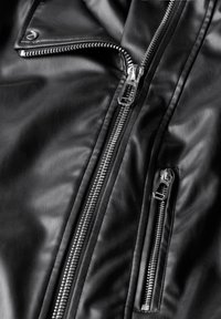 Giacca di pelle nera con numerosi zip in argento, caratterizzata da una texture liscia e elementi di design angolati, che mostrano una cucitura dettagliata.