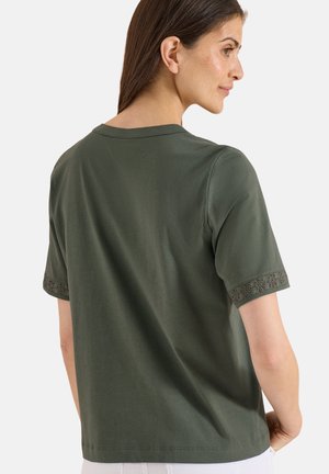 Femme portant un t-shirt vert foncé à manches courtes avec une bordure en dentelle florale sur les manches, vue de dos et légèrement tournée sur le côté.