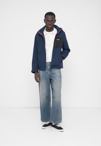 Marineblå jakke med en sort lomme, røde detaljer og hætte. Kombineret med lyseblå viddebukser og sorte sneakers.