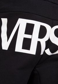 Tejido negro con letras grandes y en negrita en blanco que dicen "VERS" y "E"; textura suave, diseño casual. Vista en primer plano de la prenda.