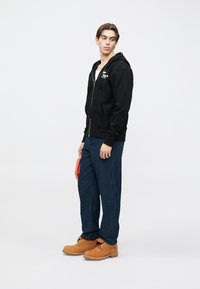 Čierna mikina na zips s predným vreckom, v kombinácii s námorníckymi nohavicami s tenkými pásmi a hnedými topánkami. Modelka drží oranžovú tašku.