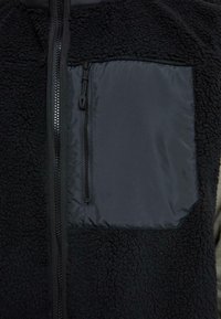 Veste polaire noire avec une fermeture éclair frontale et une poche poitrine rectangulaire avec fermeture éclair verticale.