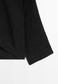 Sort tekstureret beklædningsgenstand med lange ærmer og subtile lodrette og diagonale ribbemønstre på letvægtsstof.