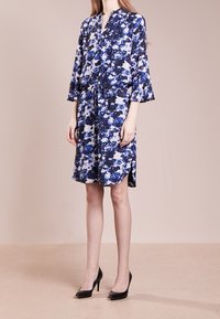 Abito floreale blu e bianco, realizzato in tessuto leggero. Presenta un colletto alla coreana, maniche a tre quarti e una vita cintrata. Tacchi alti neri.