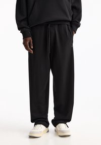 Schwarze Baumwoll-Sweatpants mit elastischem Bund und Kordelzug, ausgestattet mit seitlichen Taschen und einem dezenten Logo-Detail am Bein.