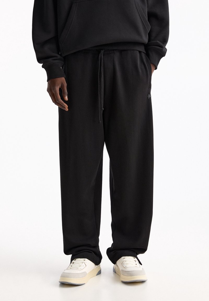 Schwarze Baumwoll-Sweatpants mit elastischem Bund und Kordelzug, ausgestattet mit seitlichen Taschen und einem dezenten Logo-Detail am Bein.