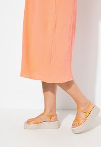 Sandalias de plataforma con una gruesa suela beige y parte superior de tiras naranja. El pie está posicionado al lado de un largo vestido melocotón texturizado.