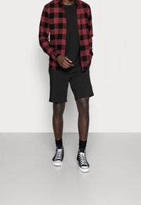 Rot-schwarz kariertes Flanellhemd über einem schwarzen T-Shirt, kombiniert mit schwarzen Shorts und hohen schwarzen Sneakers. Einfaches Design und Farben.