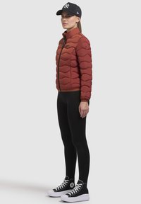 Rode gewatteerde pufferjas met hoge kraag, ritssluiting en elastische manchet, gecombineerd met zwarte leggings en zwarte hoge sneakers.