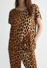 Mujer con un conjunto de top y pantalones de estampado de leopardo de manga corta, de pie con una mano en el bolsillo sobre un fondo liso.