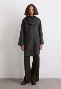 Grauer Wollmantel mit Kapuze, doppelreihigem Design, großen Vorderaschen und weiten Ärmeln. Kombiniert mit dunkelbraunen Schlaghosen und Schuhen.