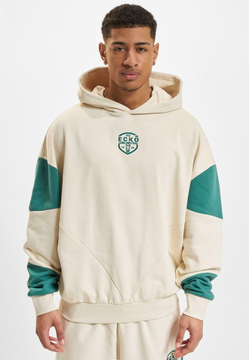 Kremet hettegenser med teal-fargede fargeblokkerte ermer, med en sirkulær grønn logo på brystet og ribbestrikkede mansjetter. Glatt tekstur, uformell design.