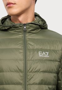 Grønn nylon puffjakke med glidelås, med horisontale sømmer og hette. Brodert logo på brystet. Glatt tekstur.