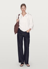 Bílá volná košile s přeloženým límcem a tříčtvrtečními rukávy, v kombinaci s tmavými džínovými kalhotami s širokými nohavicemi a černými sandály. Hnědá tkaná kabelka.