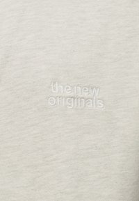 Gaiši pelēks audums ar mazu baltu izšūtu tekstu "the new originals", kas ir izvietots centrā.