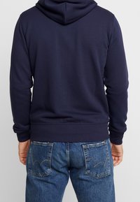 Sweat à capuche bleu marine avec une poche kangourou et des poignets côtelés, associé à un jean en denim avec un design distinctif sur la poche arrière.