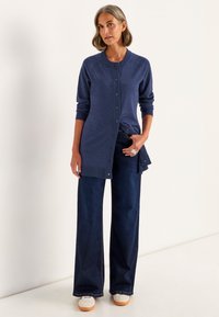 Frau in einem marineblauen, langen, geknöpften Cardigan, mit weiten dunklen Jeans, weißen Sneakern und einem silbernen Statement-Ring, der an einer weißen Wand steht.