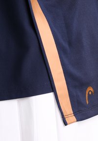 Marinblå sporttröja med strukturerad tyg, en orange sidosten och en logotypdetalj nära fållen. Fållen är rak och välutförd.