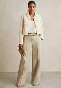 Beige cropped jas, geweven stof met twee voorzakken; witte gebreide top; brede tankleurige broek met plooien; tankleurige clutch met franje-details.