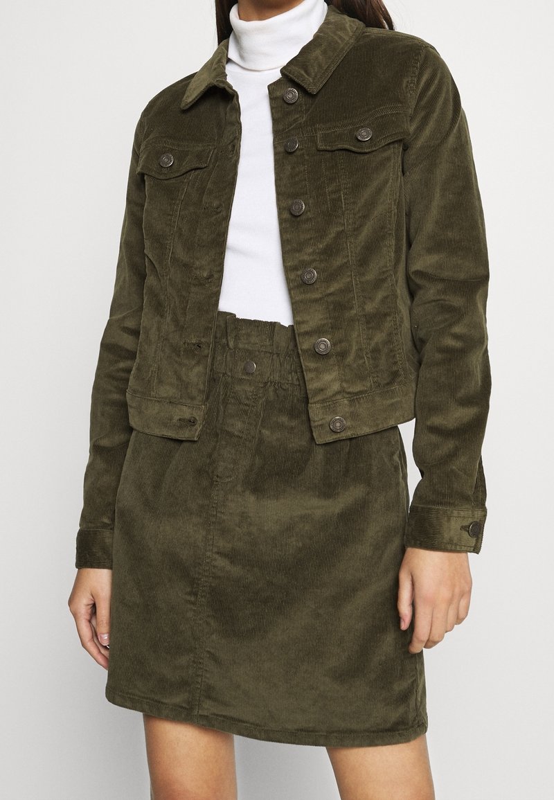 Woman wearing olive green corduroy jacket and matching skirt over a white turtleneck, standing against a plain light background.