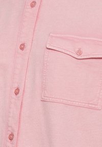 Lichtroze knoopshirts met een zak, voorzien van bijpassende roze knopen en een gestructureerde afwerking. De stof lijkt zacht en casual.