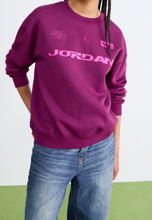 Personne portant un sweat Jordan violet avec des logos et du texte roses, associé à un jean en denim bleu, debout avec une main derrière le dos.