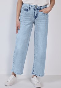 Jean large bleu clair en denim, avec taille haute, décoloration subtile et design classique à cinq poches, associé à des baskets blanches.