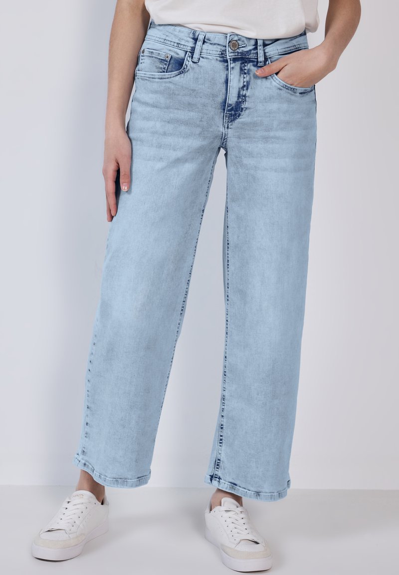 Jean large bleu clair en denim, avec taille haute, décoloration subtile et design classique à cinq poches, associé à des baskets blanches.