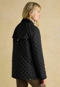 Geacă neagră matlasată cu guler, având cusături în formă de diamante și o margine curbată la spate, purtată peste pantaloni în carouri.