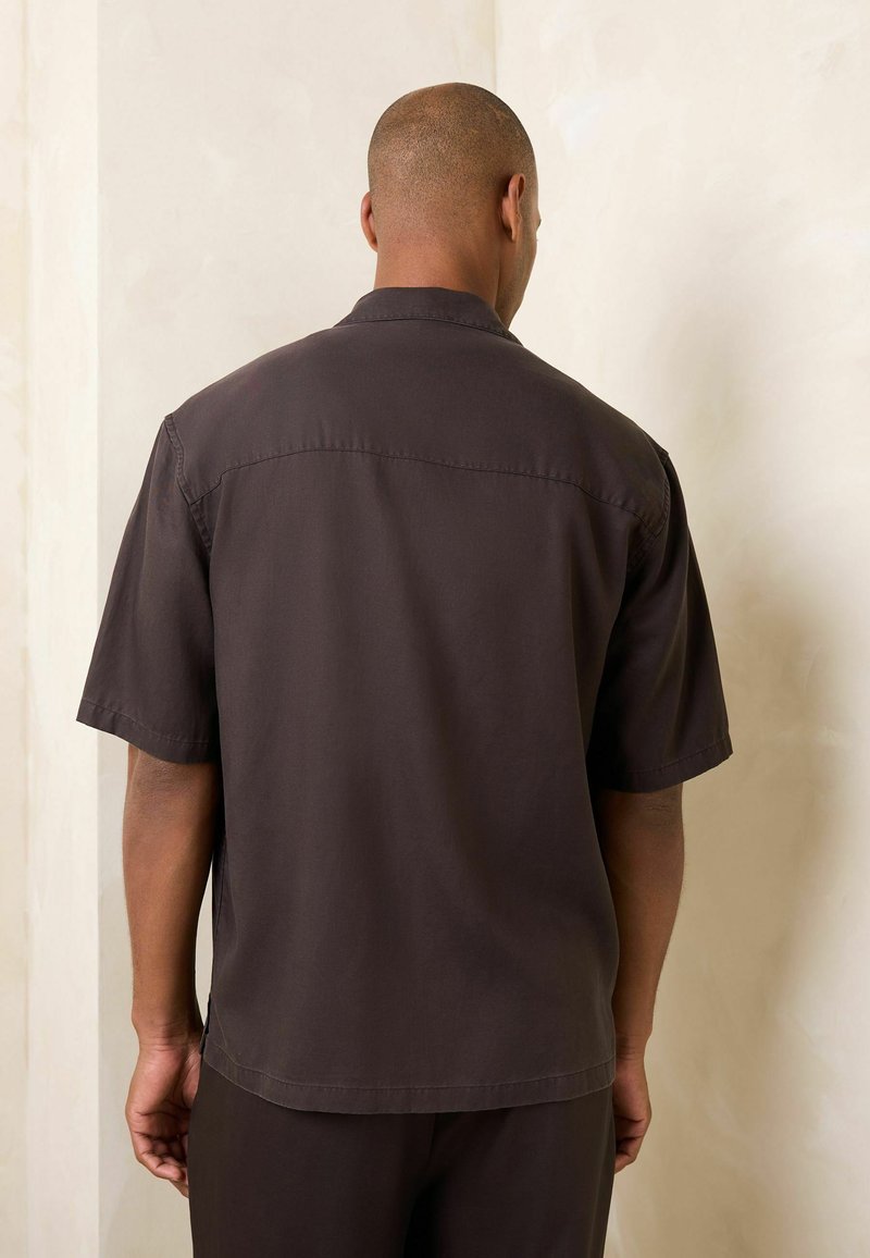 Homme de dos portant une chemise sombre à manches courtes avec col et un pantalon assorti sur un fond clair uni.