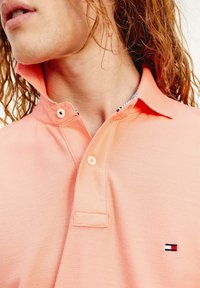 Kortärmad pikétröja i ljus persikoröd, med en texturerad tyg, tvåknappsknappning och en liten broderad logotyp på bröstet.