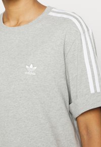 Person som bär en ljusgrå Adidas-t-shirt med vita tre ränder på axeln och Adidas-logotypen på bröstet.
