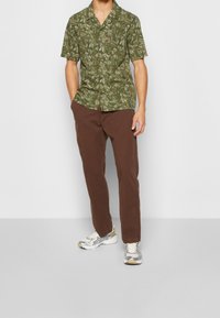 Man som bär en grön kortärmad skjorta med lövdesign, bruna byxor och vita och silverfärgade sneakers, stående mot en enkel bakgrund.