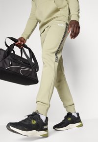 Un pantalon de jogging vert olive clair présente un design fuselé avec une bande logo noire. Des chaussures de sport noires et un sac de sport noir avec des accents blancs complètent le look.