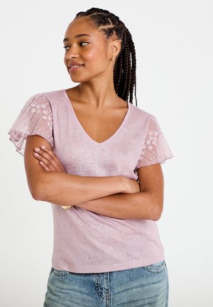 Femme aux cheveux tressés portant un haut rose pâle à col en V avec des manches courtes transparentes brodées de fleurs, bras croisés, regardant sur le côté.