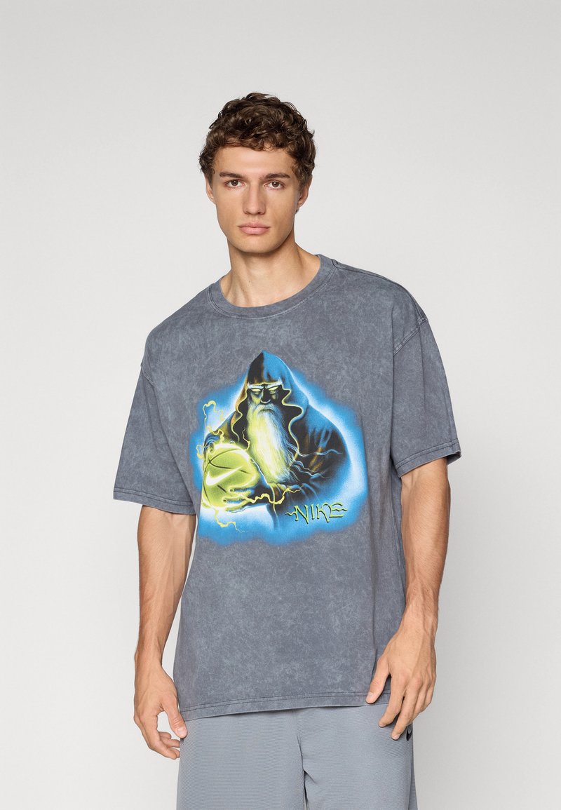 Junger Mann in einem grauen T-Shirt mit einem grafischen Druck eines Zauberers, der einen leuchtenden Basketball hält, und dem Wort "Nike" in Neonfarben.