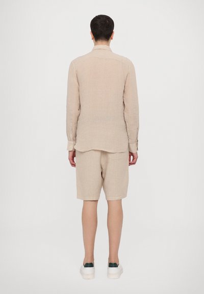 Camisa de linho bege de manga longa combinada com calções a condizer, apresentando um corte relaxado, botões e uma textura suave. Sapatos brancos com detalhes verdes.