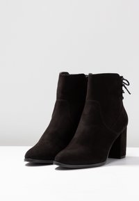 Un paio di stivaletti neri in suede con tacco a blocco e dettaglio di lacci sul retro, su una superficie bianca e sfondo grigio.