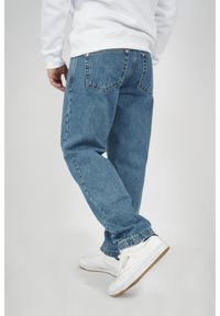 Person klädd i blå jeans, vit sweatshirt och vita sneakers som kliver framåt mot en enkel ljus bakgrund.