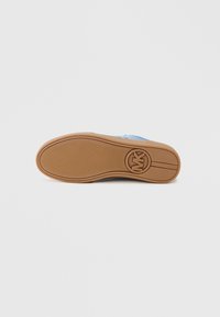 Niebieski trampek z teksturowaną tkaniną wierzchnią, gumową podeszwą i okrągłym logo na podeszwie. Podeszwa ma wzorzysty bieżnik zapewniający przyczepność.