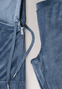 Pantalon et veste en tissu doux bleu avec fermeture éclair et cordons, posés à plat sur un fond clair, montrant la texture et les détails du matériau.