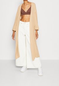 Beżowy długi kardigan z balonowymi rękawami, brązowy biustonosz i białe szerokie spodnie połączone z białymi trampkami. Gładki materiał, luźny krój.