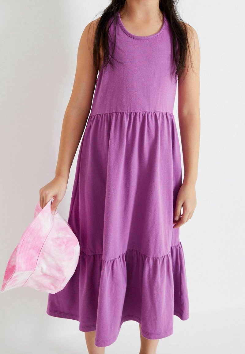 Jeune fille portant une robe violette sans manches, tenant un chapeau bob rose tie-dye sur un fond blanc uni.