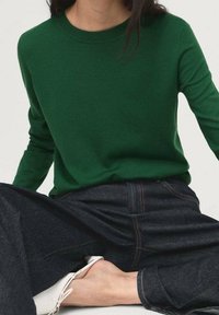 Personne portant un pull vert à manches longues, un jean foncé et des chaussures blanches assise en tailleur devant un fond uni.
