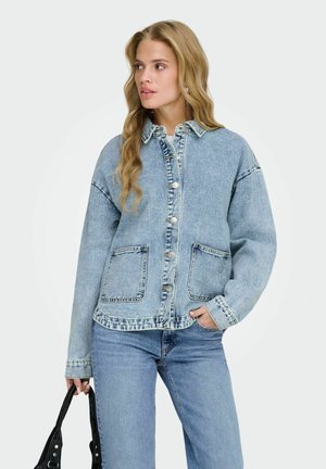 Hosszú szőke hajú nő világoskék farmer dzsekit és farmert visel, fekete kézitáskát tart, és oldalra néz.