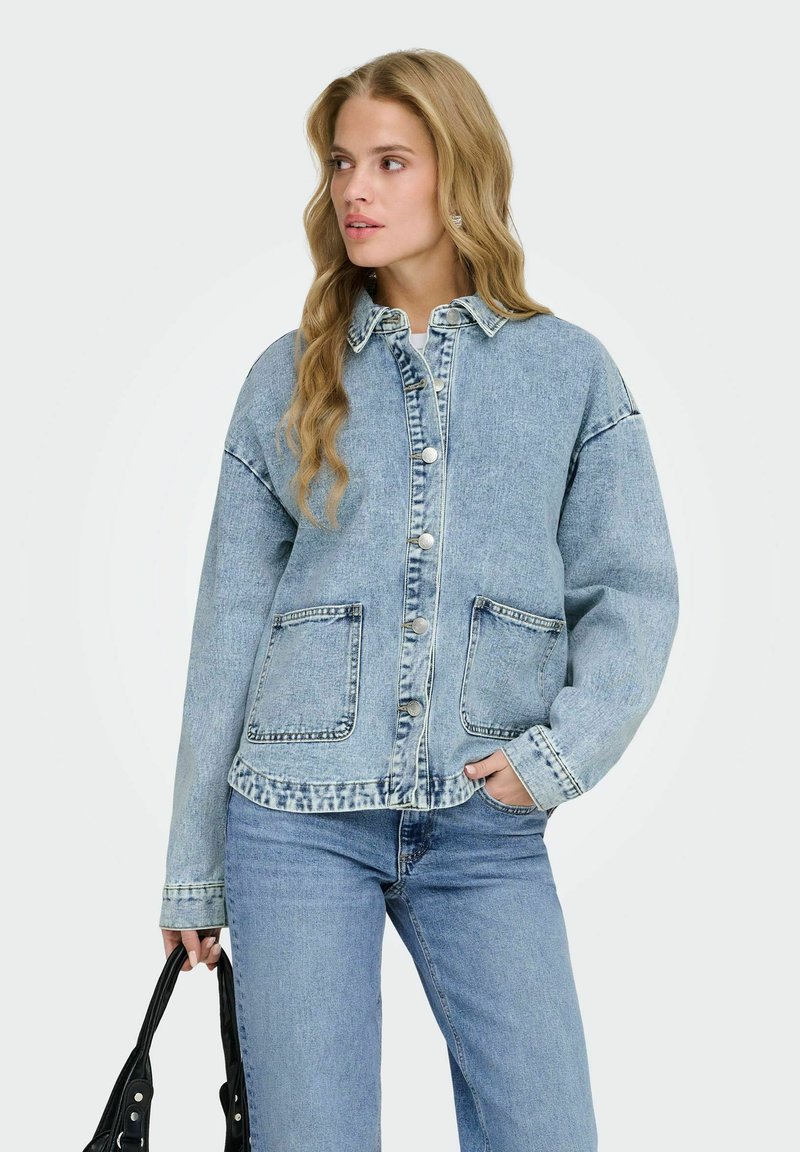Femme aux longs cheveux blonds portant une veste en jean bleu clair et un jean, tenant un sac à main noir, regardant sur le côté.
