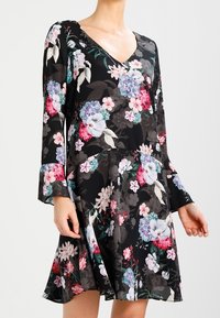 Mustan kukallisen mekon, jossa on V-mallinen kaula-aukko, pitkät kellotaskut ja liehuva muotoilu. Kangas esittää monivärisiä, yksityiskohtaisia kukkakuoseja.