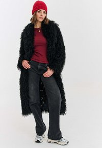 Černý huňatý kabát z umělé kožešiny, vínový přiléhavý top, tmavé džíny a bílé tenisky. Červená pletená čepice doplňuje vzhled.