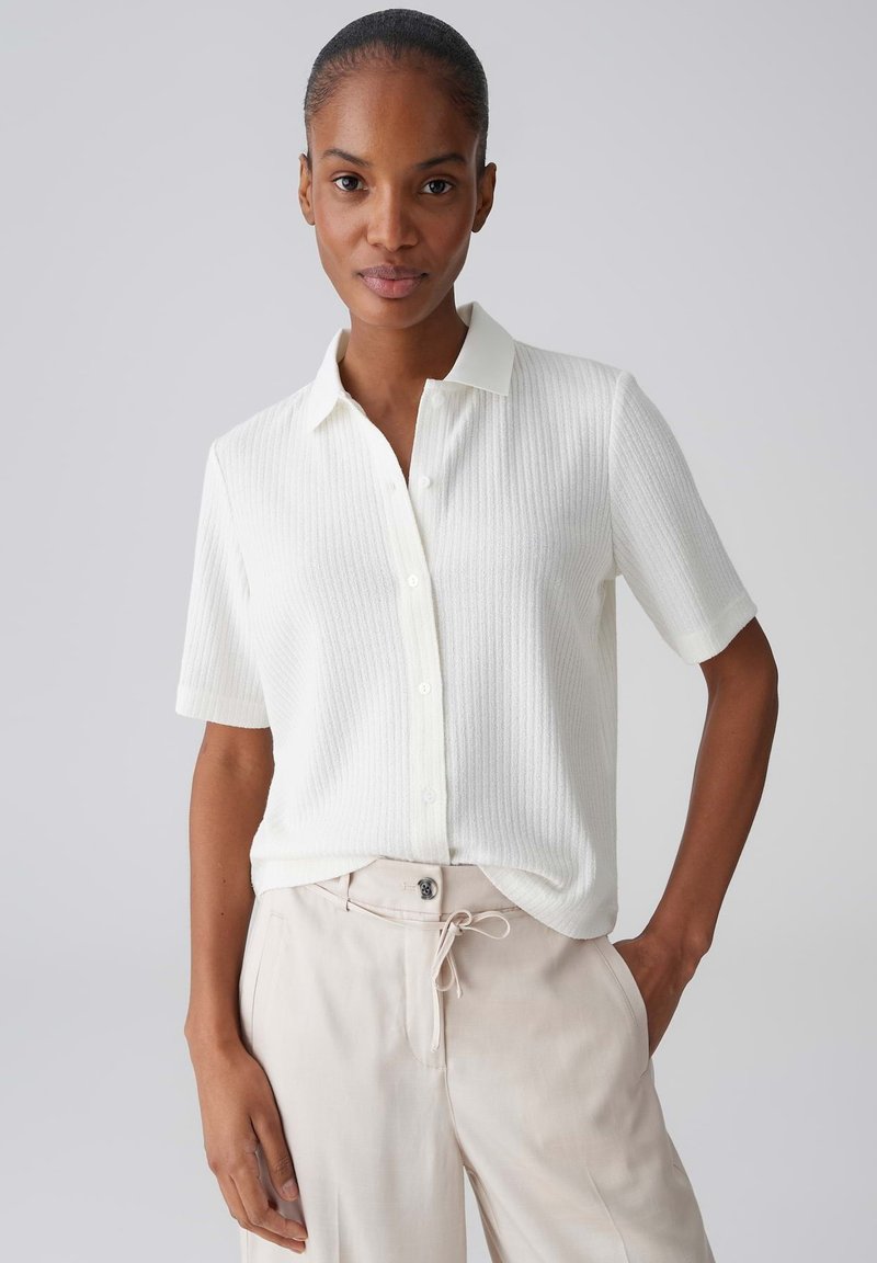 Donna che indossa una camicia bianca a maniche corte con bottoni e tessuto strutturato, infilata in pantaloni beige con coulisse, in piedi con una mano in tasca.