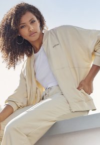 Beige Sport-Jacke mit Reißverschluss und gerippten Bündchen, kombiniert mit einem weißen, taillierten Shirt und passendem beigen Jogginghose. Mit gesticktem Logodetail.